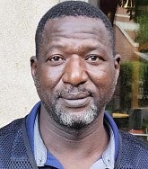African man with greying beard and short hair looks calmly at the camera; he is wearing a blue collared short and a UO Facilities Services vest