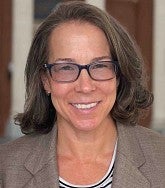 White woman with shoulder length brown hair, dark blue- rimmed glasses, ear rings, brown jacket, and striped shirt smiles at the camera.