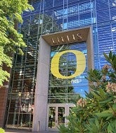 Glass-fronted building with a large yellow O and the word LILLIS in concrete over the O. Trees and bushes on left and right of the building.