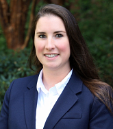 White woman with long brown hair, wearing a white collared shirt and blue blazer smiles at the camera