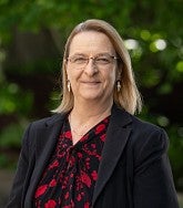 White woman with shoulder length blonde hair, glasses, earrings, black jacket, and red-and-black top. Greenery behind her.