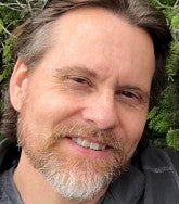 White man with brown hair and greying beard smiles at the camera; greenery behind him