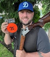 White man in a Dodgers cap holding a skeet shooting rifle; he also holds an orange "25" disc indicating how many clay skeet targets he shot in a row