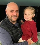 Bald white male with stubble and moustache holding his 6-year-old daughter who is smiling in a red dress