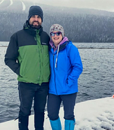 White man and woman wearing woolly hats and winter clothing stand on snow next to a dark lake; trees in background