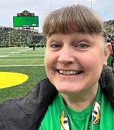 White woman with brown hair tied back; standing on the field at Autzen stadium; wearing a green UO Ducks shirt and black jacket