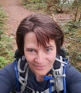 White woman with short brown hair wearing a blue shirt and hydration backpack; trail and trees in background
