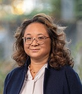 Woman with shoulder-length wavy brown hair wearing glasses, a navy blazer and white shirt smiles for the camera; greenery in the background