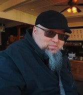 Asian-American man with a stylish grey beard in a black ball cap and sunglasses looks into the camera; coffee shop in the background