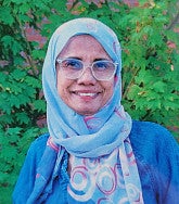 Woman wearing glasses, a headscarf, and blue top smiles at the camera; a red brick building and green foliage are visible behind her 