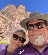 Gary wearing a hat and sunglasses (right) with his wife Kathy (also in sunglasses) at Zion National Park  - sandstone cliffs and blue sky behind them
