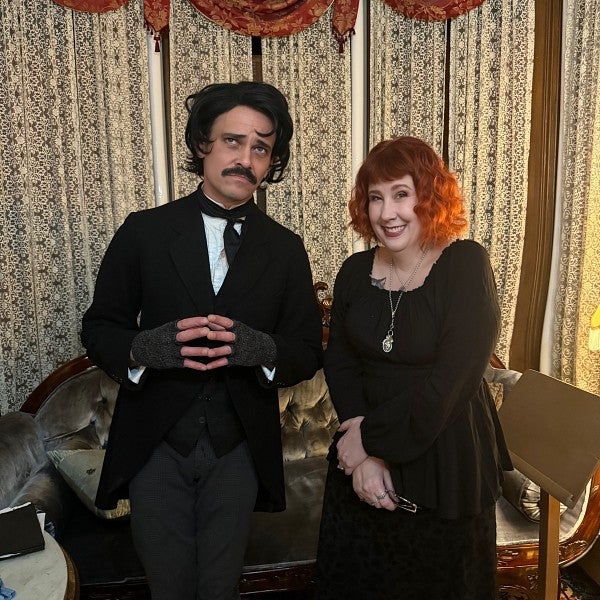Tall white man with black hair and a mustache, in black jacket and grey trousers; next to a white woman with red hair and a black dress; beige drapes in the background