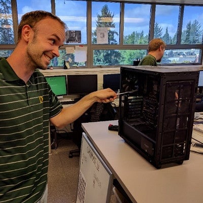 Man with short hair and mischievous grin prods a screwdriver into a desktop computer tower