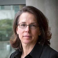 A woman with brown shoulder-length hair glasses and a dark shirt looks warmly at the camera