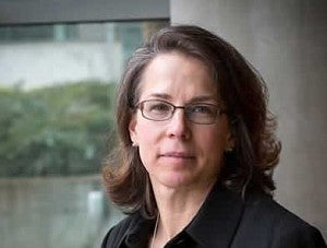 A woman with brown shoulder-length hair glasses and a dark shirt looks warmly at the camera