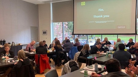 Screen showing 'Fin' and 'Thank you'. 20 LEADS participants seated around tables taking notes and discussing process improvement; pink flowering tree visible outside through window