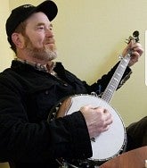 Side view of a man wearing a baseball hat and black jacket playing a five-string banjo