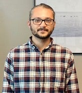 Man with glasses, a moustache and closely cropped beard, and a checkered shirt looks straight at the camera