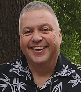 Man with short grey hair and dark blue shirt smiles broadly in front of greenery