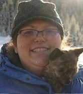 Woman in glasses and woolen hat; dog peeking out of her blue jacket; snowy landscape in background