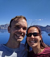 Man with short brown hair and woman with shoulder-length brown hair smile in front of Crater Lake (blue sky, blue water, crater edge in background)
