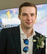 Young white man with short blond hair, blue eyes, collared white shirt, black jacket and sunglasses on his shirt; corsage on his right lapel