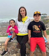 Woman with long dark hair and olive skin sits on a low wall with her 6-year-old daughter at left and 11-year-old son at right; sea and sand visible behind them