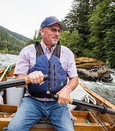 Man with a ball cap and glasses rows a boat in a river; raging waters and green trees in the background. 
