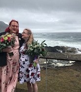 A shorter woman with long blond hair looks up at a taller woman with shoulder-length red hair; both are wearing glasses and holding bouquets; behind them is a beautiful coastal scene