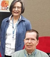 Woman with short hair, glasses, a white shirt and blue jean jacket stands smiling at left; at right seated next to her is a man with a moustache