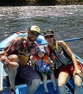 A man and a woman in sunglasses, shirts, shorts, lifejackets and caps sit in an inflatable boat on the river; between them is a small child with a lifejacket; all are looking at the camera; a paddle is behind them