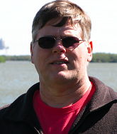 Man with sunglasses, red shirt, and brown Columbia sweatshirt in the sun with body of water behind