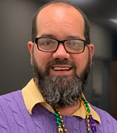 Bearded man in black rimmed-glasses looks cheerily at the camera; he is wearing a purple sweater and green-gold-purple beads
