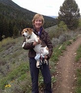Woman in a brown coat smiles on the side of a hiking trail, a medium brown-and-white dog in her arms